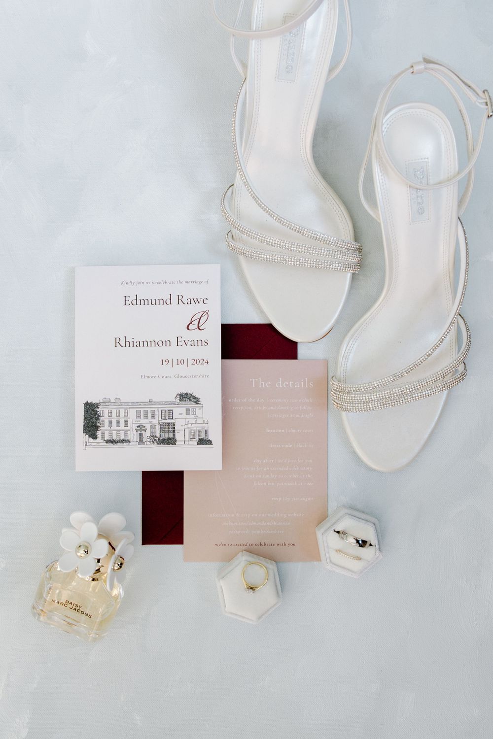 Elegant wedding invitation suite with white strappy heels and perfume bottles on light background.