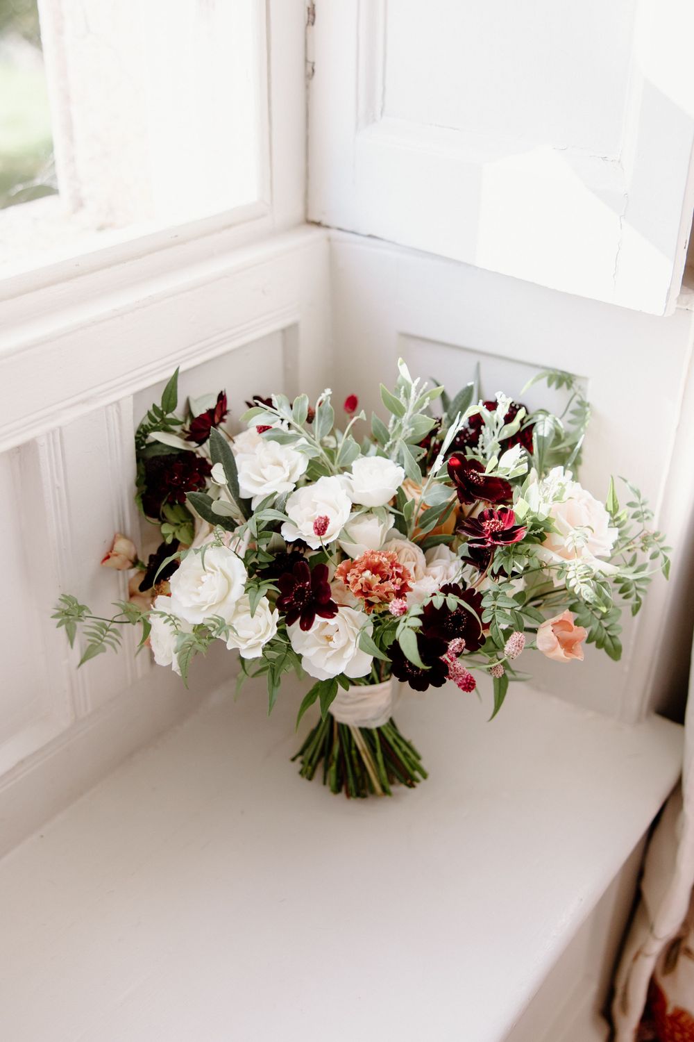 Stunning bridal bouquet with white roses and deep burgundy flowers on a windowsill.