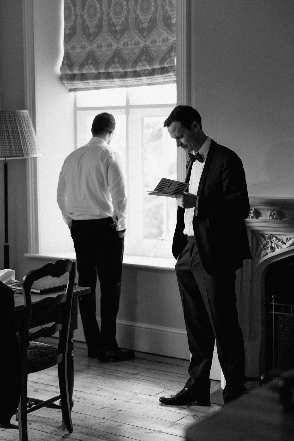 Two men in formal attire stand in a sunlit room as one reads from a document.