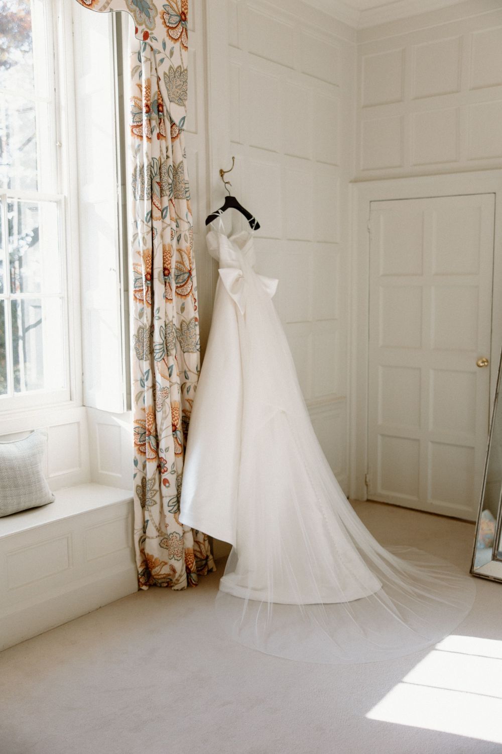 An elegant white wedding dress hanging in a bright, classic room with floral curtains.