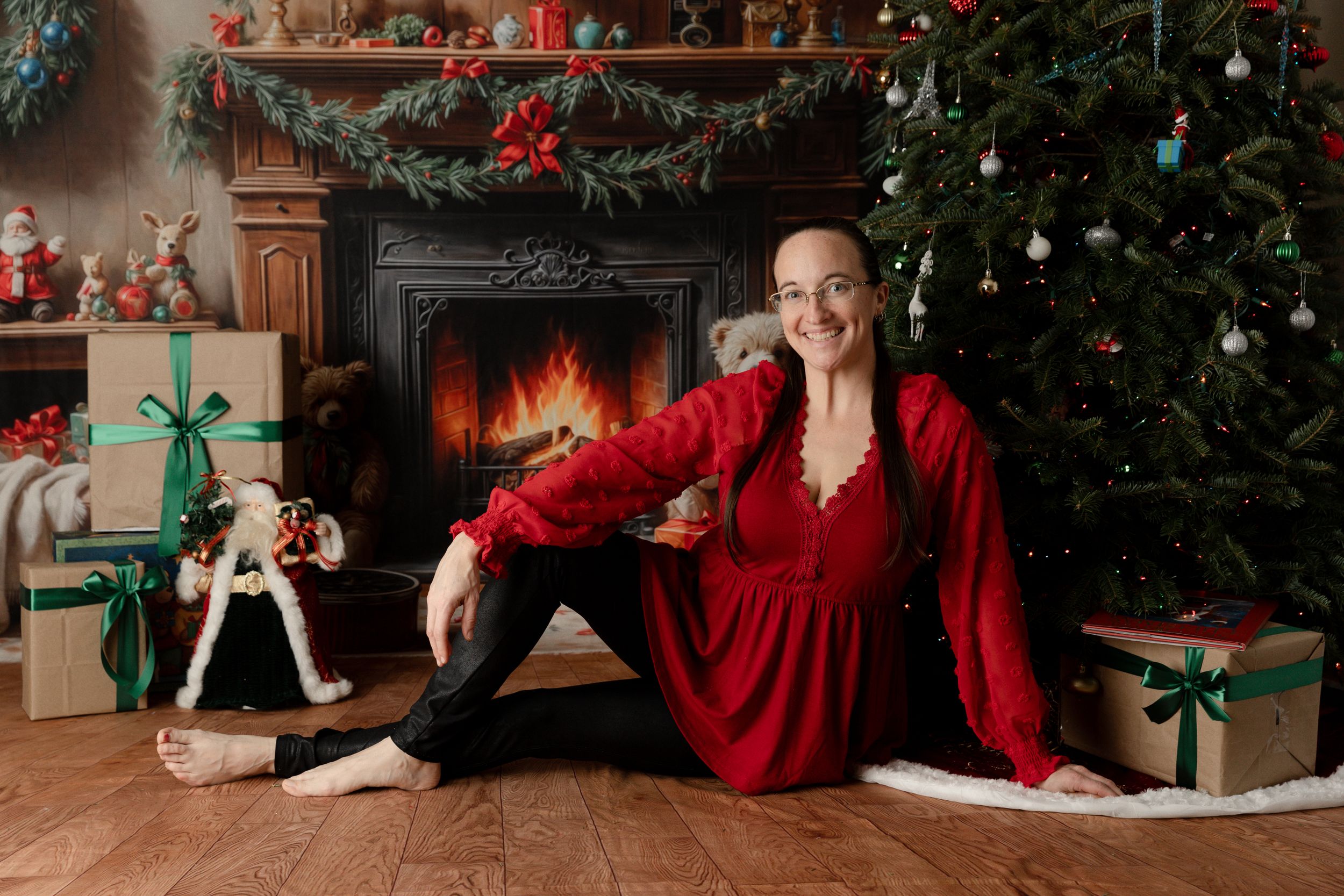 Smiling woman in red dress sitting by decorated Christmas tree and fireplace with holiday gifts.