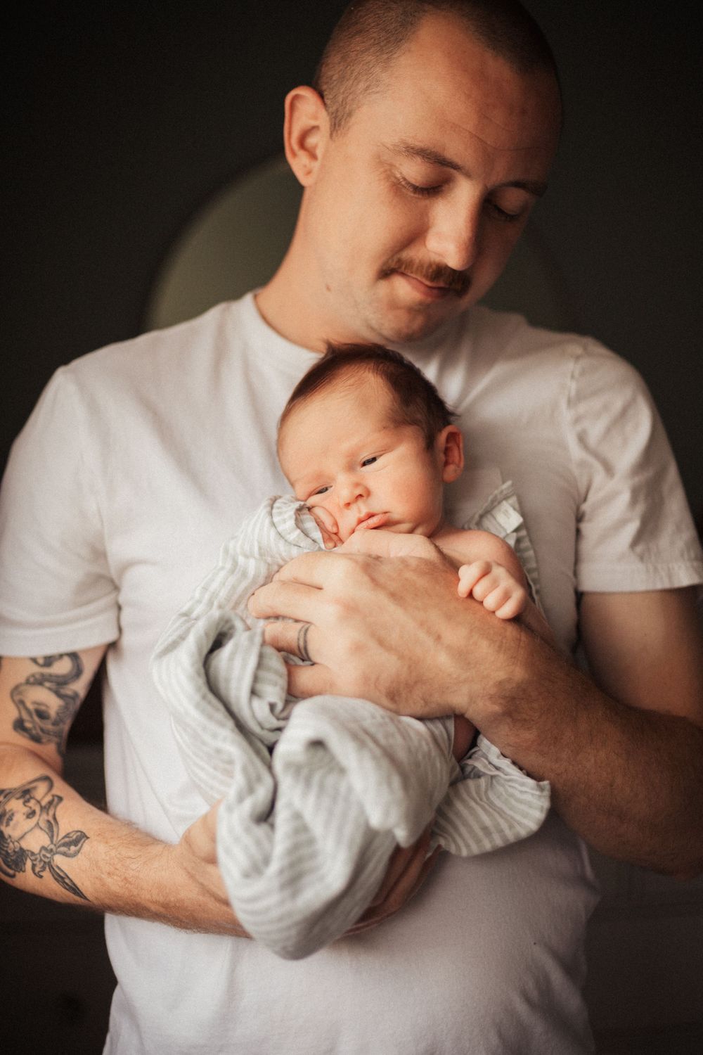 A tattooed parent in white shirt cradles sleeping newborn wrapped in light gray.