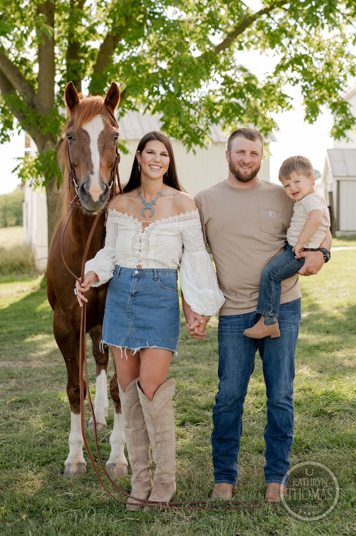 Family with horse poses together in rustic farm setting under large tree.