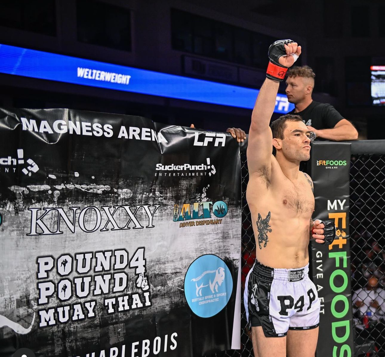 A victorious MMA fighter raises his arm in celebration inside an octagon cage during a Muay Thai event.