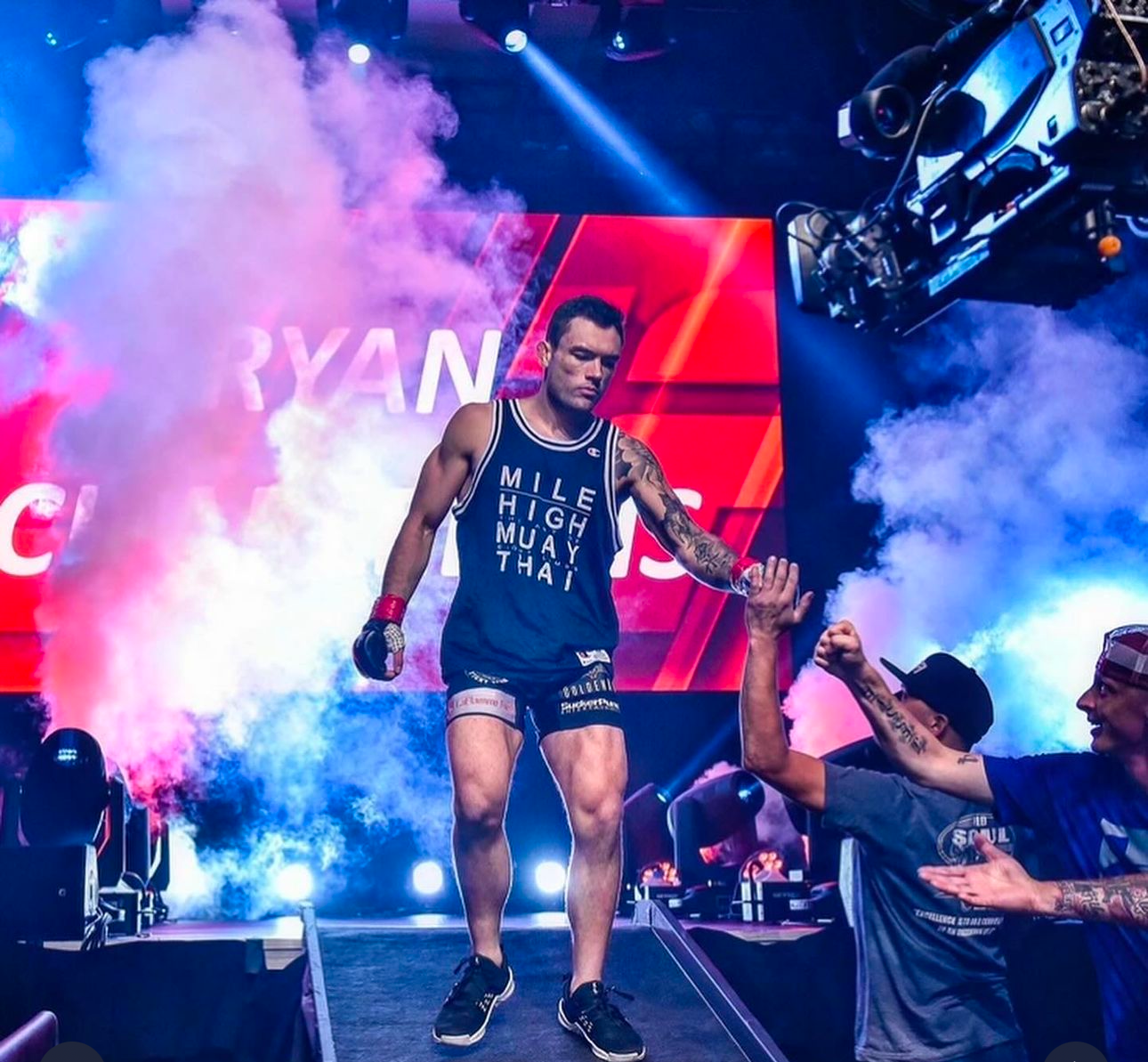 Professional wrestler makes dramatic entrance on stage with blue spotlights and smoke effects during wrestling event.
