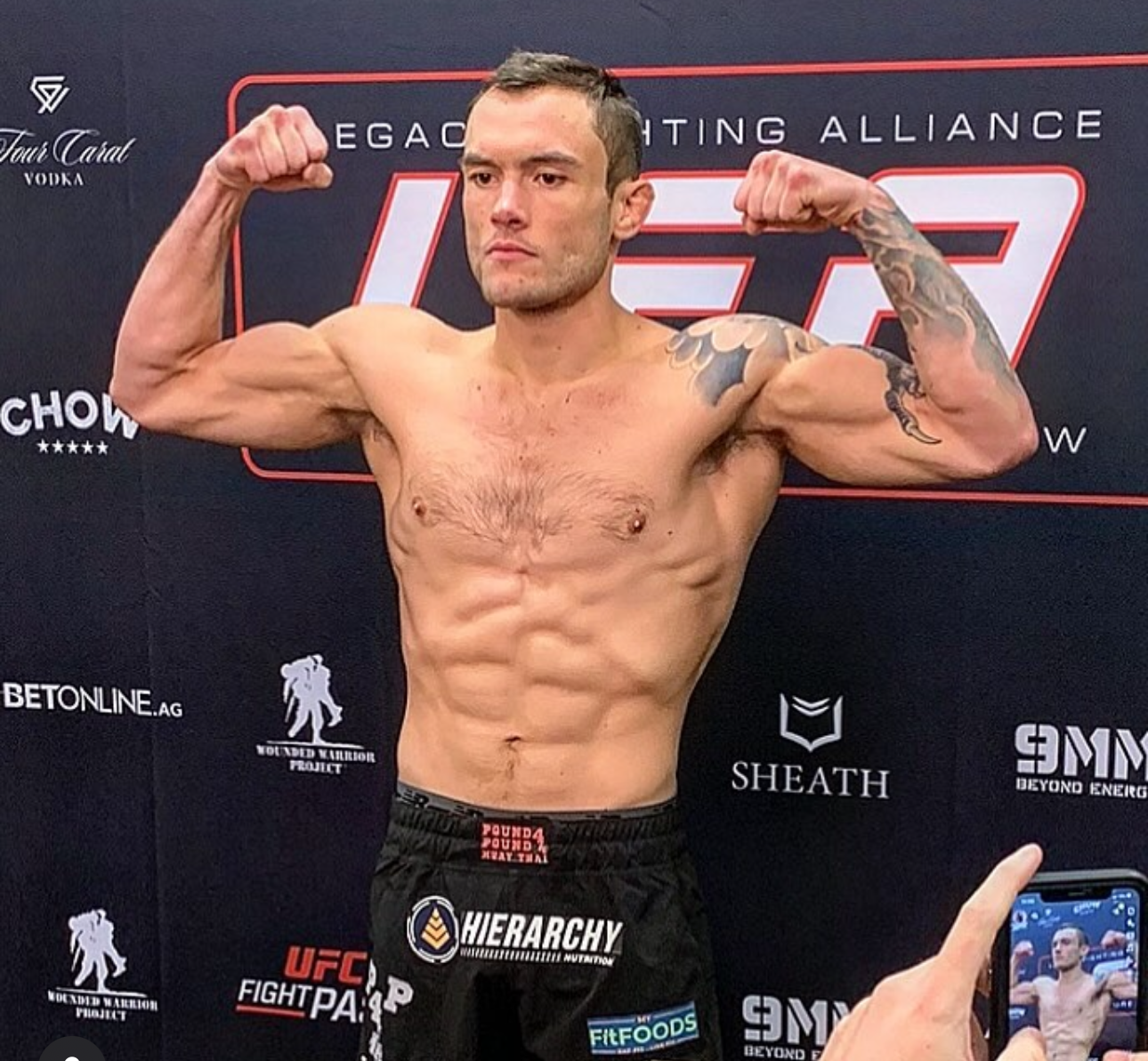 Shirtless MMA fighter flexes muscles and poses during weigh-in event with sponsor banners visible in background.