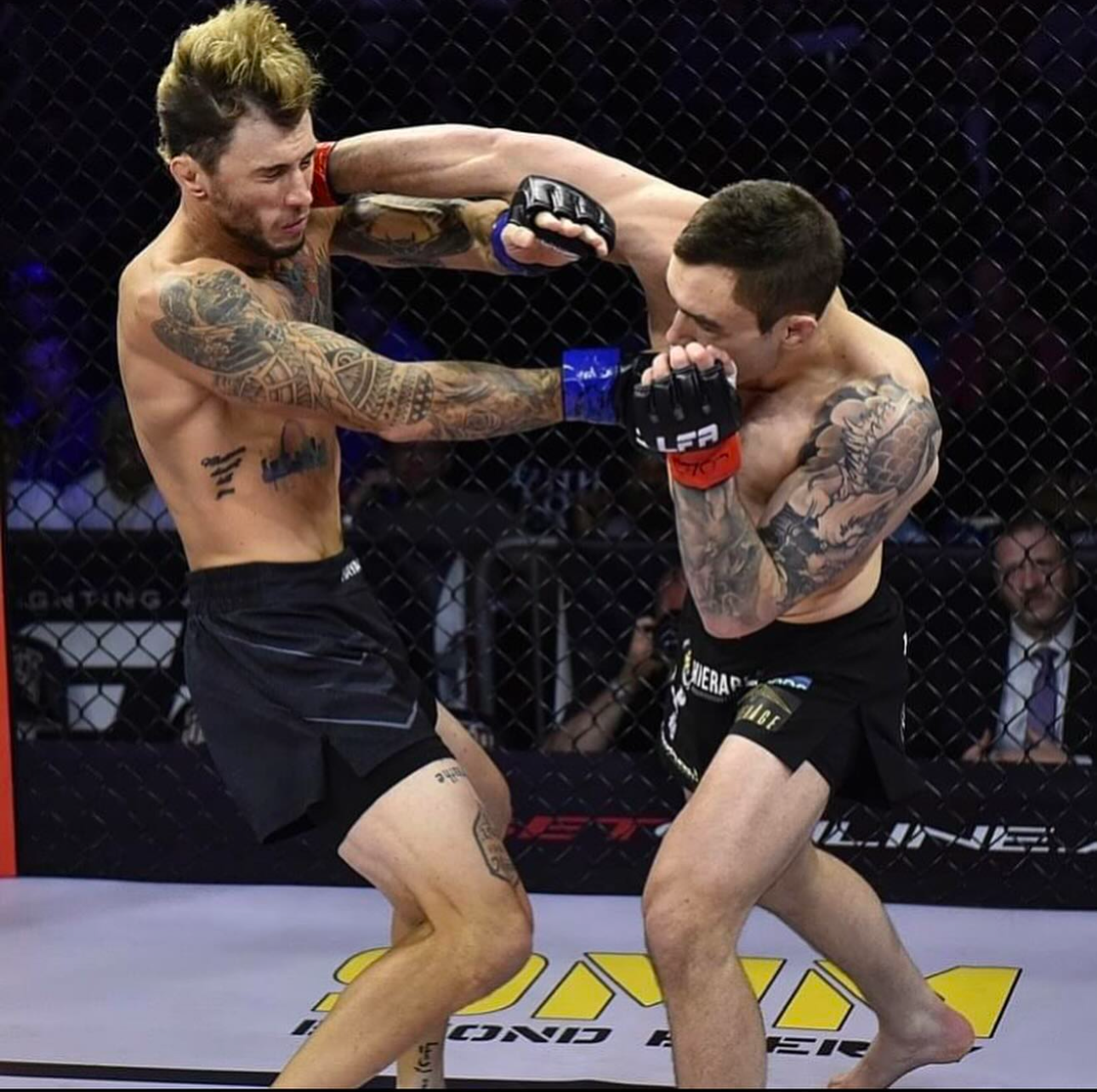 Two MMA fighters exchange strikes during an intense cage match with visible tattoos on both competitors.