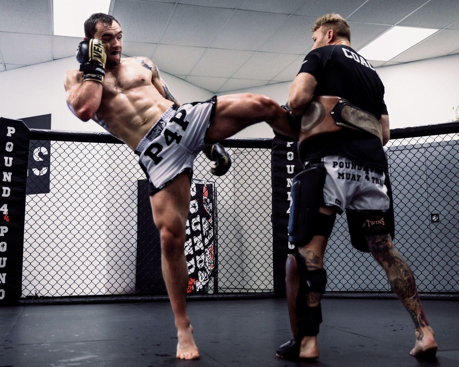 Two MMA fighters sparring in an octagon cage, with one executing a high kick during training.