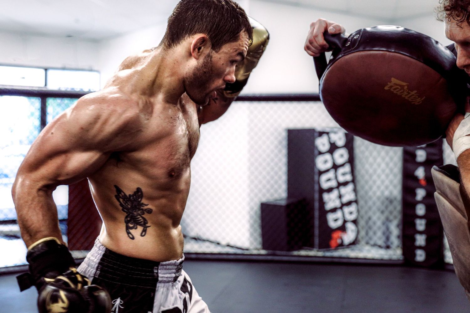 Shirtless fighter with tattoo and boxing gloves training with medicine ball in MMA gym.