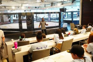 Students attend a lecture in a modern classroom with long white desks and industrial-style exposed ceiling.