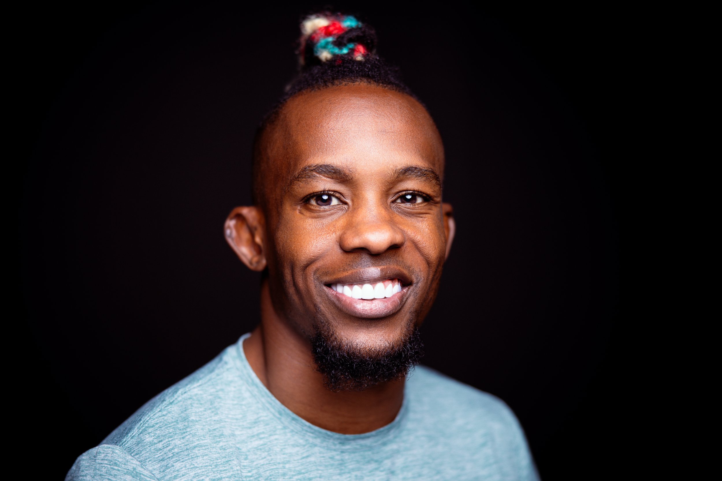 Smiling person wearing light blue t-shirt and colorful hair accessory against black background.
