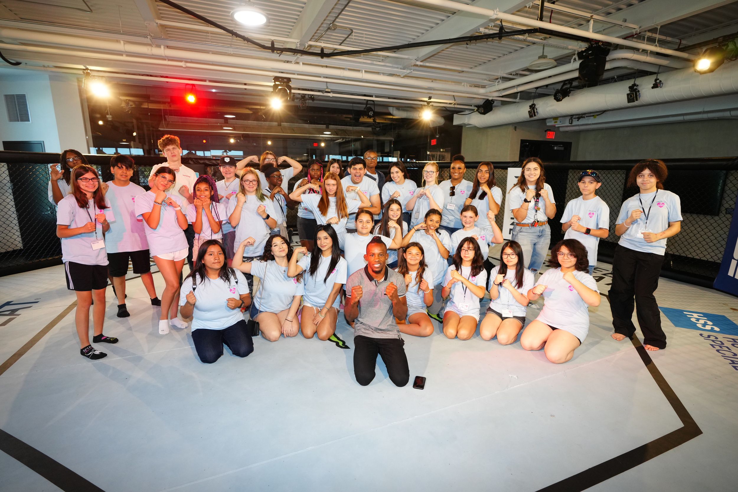 A large group poses together in white t-shirts at an indoor venue with stage lighting.