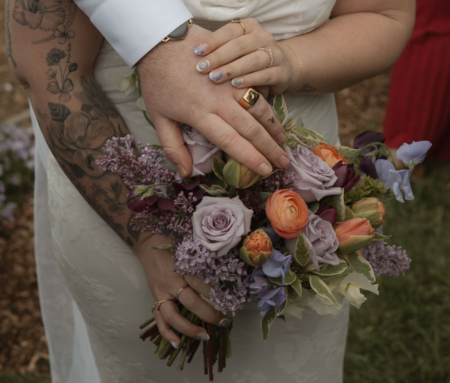 A beautiful bouquet of pink roses, orange ranunculus, and lilac flowers held by hands with floral tattoos and wedding rings.