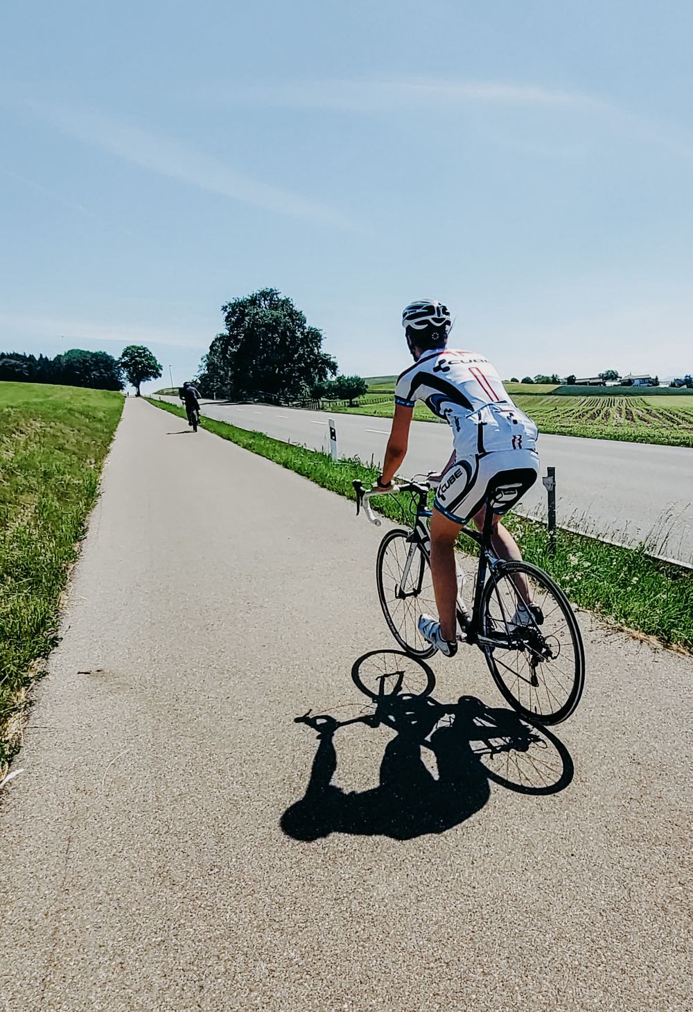 Road cyclist in white jersey riding narrow country road on sunny day.