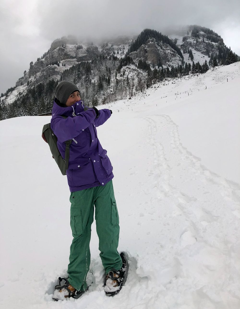 Person in purple jacket and green pants snowshoeing through snowy mountain landscape with misty peaks in background.