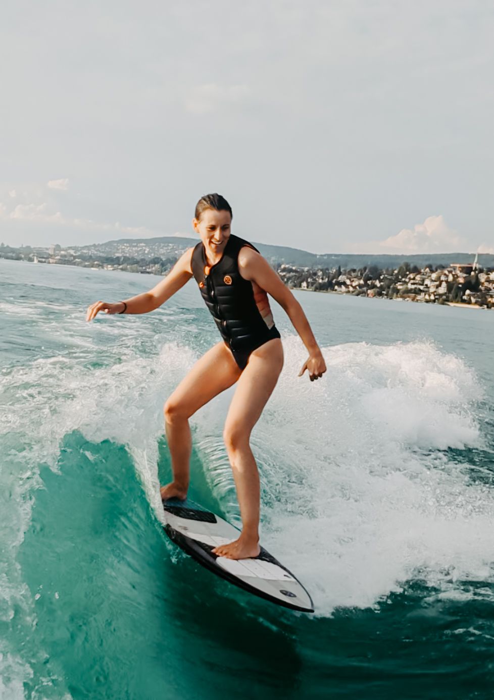 Young woman in black swimsuit surfing a wave on turquoise water with a coastal town in the background.
