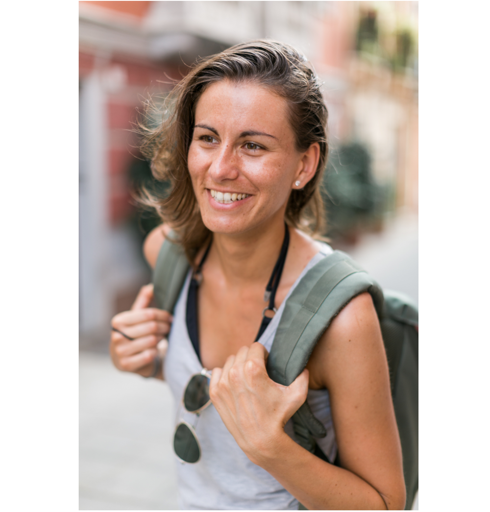 Smiling young woman with backpack and sunglasses exploring a European city street on a sunny day.