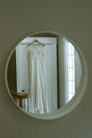 A white wedding gown with delicate lace details hangs on a wooden hanger, reflected in a round wall mirror.