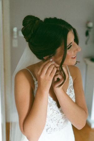 A bride in a white lace dress puts on earrings while smiling and wearing a delicate veil.