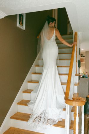 Bride in elegant white mermaid wedding dress with long train ascending wooden staircase in a home.