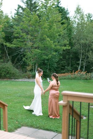 Two wedding attendees in elegant dresses walk together across a lush green lawn surrounded by evergreen trees.