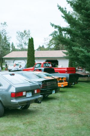 Classic cars parked in a row on grass with a white house and evergreen trees in the background.