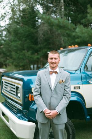 A person in a light grey suit and brown bowtie poses in front of a vintage blue truck at an outdoor event.