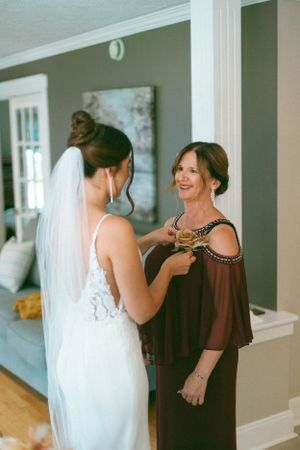 A bride in a white lace dress getting ready with help from someone wearing a brown flowing dress.