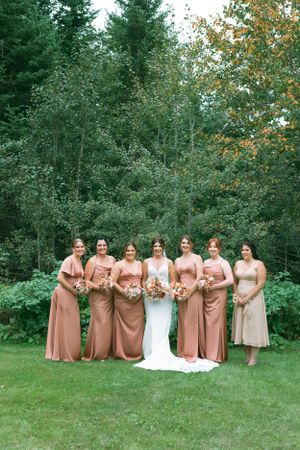 A wedding party standing together outdoors in dusty rose colored dresses against a backdrop of evergreen trees.