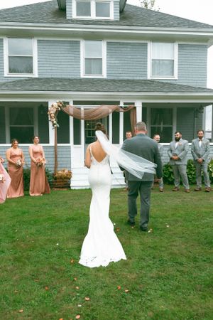 Wedding party gathers on the lawn of a blue shingled house during an outdoor ceremony.