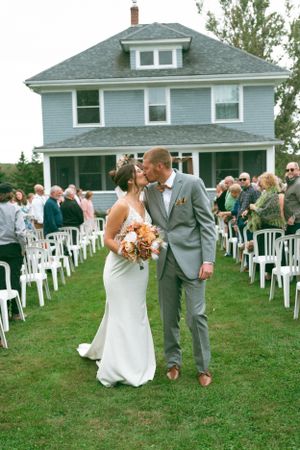 An outdoor wedding ceremony on a lawn with white chairs and a blue house in the background.