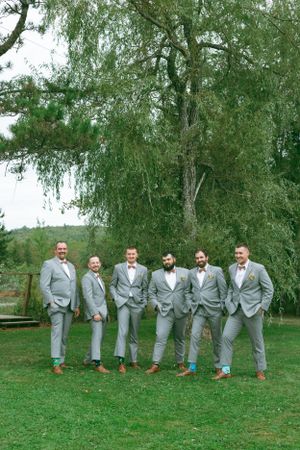 Groomsmen lined up outdoors wearing matching light gray suits under a large weeping willow tree.