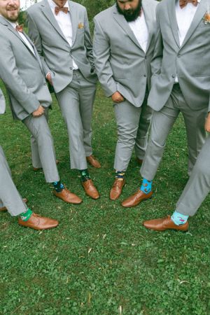 Groomsmen in matching gray suits show off their brown dress shoes and colorful turquoise socks on grass.