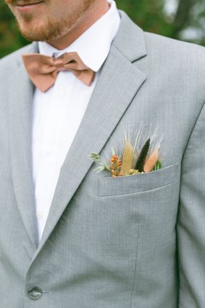Close up of a light gray suit jacket with peach and feather boutonniere and brown bowtie for a wedding.