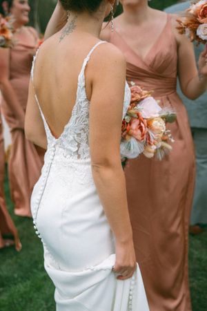 A bride in a white lace dress and bridesmaids in peach gowns holding coral and blush floral bouquets at an outdoor wedding.