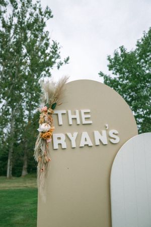 A decorative wedding sign reading 'THE RYANS' adorned with dried flowers and pampas grass against a green backdrop.