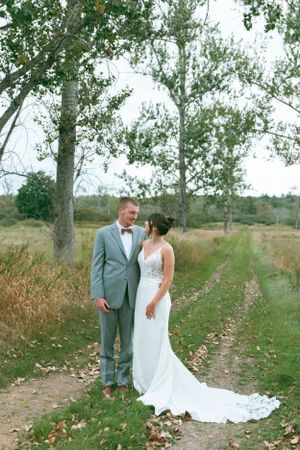 A couple in wedding attire stands on a dirt path surrounded by autumn trees and fallen leaves.