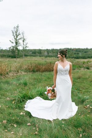A bride in a fitted lace gown holds autumn florals while standing in a scenic field landscape.