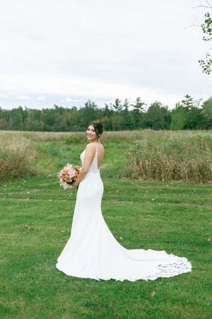 A stunning white wedding dress with a long train is showcased in an outdoor field setting with trees in the background.