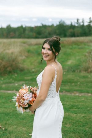 A bride in a fitted white lace dress holds a peach-colored bouquet while standing in a grassy field with trees in background.
