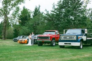 Classic cars including a red pickup truck and other vintage vehicles parked on grass with evergreen trees in background.