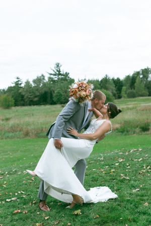 Couple shares romantic kiss in grey suit and white wedding dress during outdoor countryside celebration.