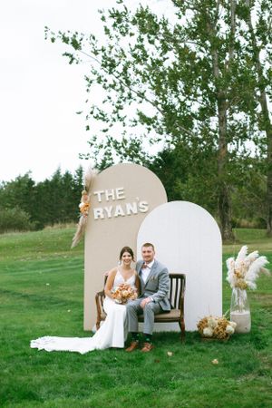 A wedding setup outdoors with an elegant curved white and beige backdrop and bench against a green natural background.