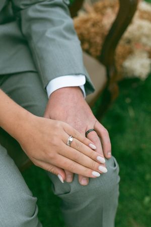 Close-up of hands wearing wedding rings resting on a grey suit pant leg showing elegant manicured nails.