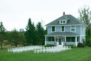 A two-story white farmhouse stands behind a large display of small white lawn decorations arranged in rows on green grass.