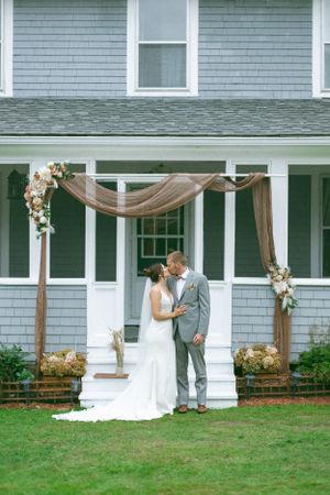 A decorated wooden arch with tan fabric and floral accents stands on the porch of a gray shingled house.