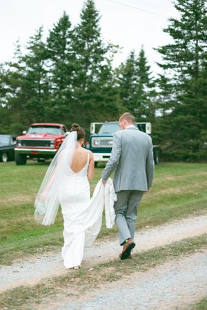 Wedding couple walks down a gravel path beside vintage trucks and evergreen trees.