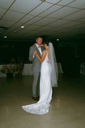 A couple shares their first dance at a wedding reception in a dimly lit banquet hall with white ceiling tiles.