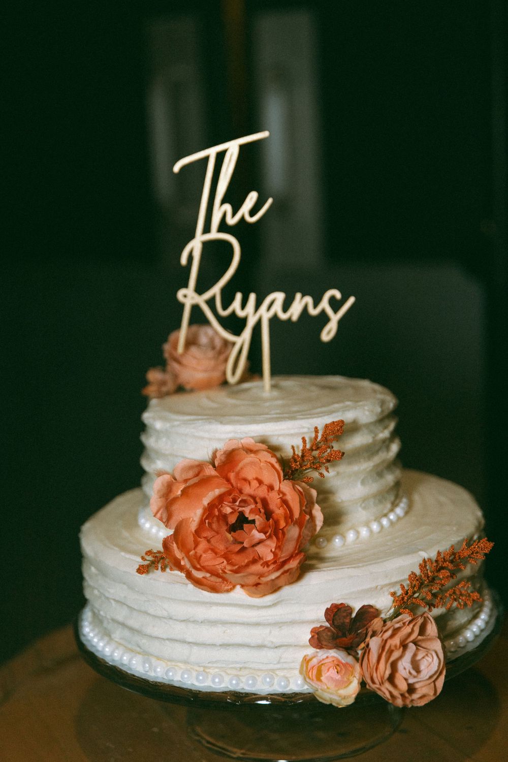 White tiered wedding cake decorated with peach-colored roses and a custom 'The Ryans' topper against dark background.