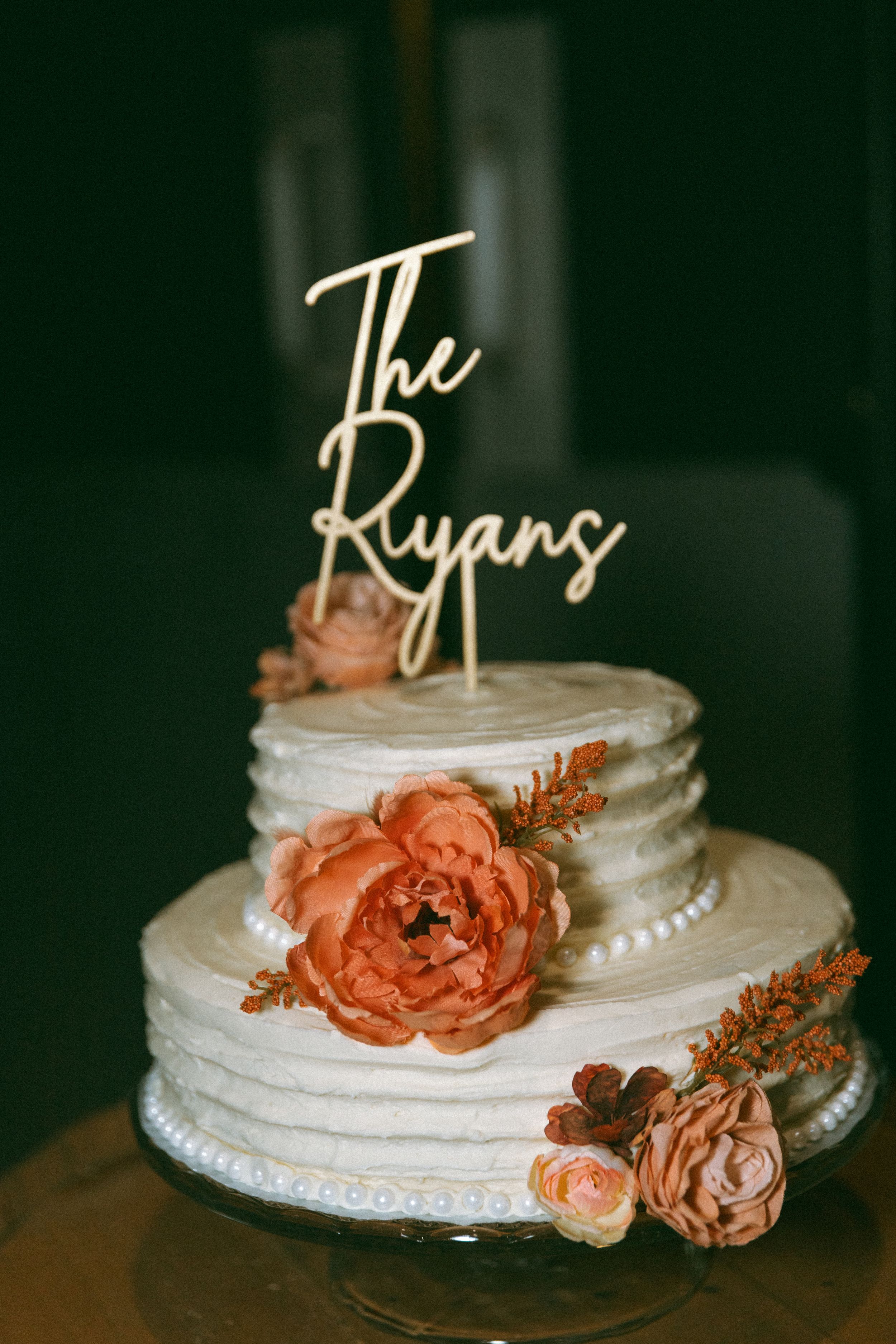 White tiered wedding cake decorated with peach-colored roses and a custom 'The Ryans' topper against dark background.